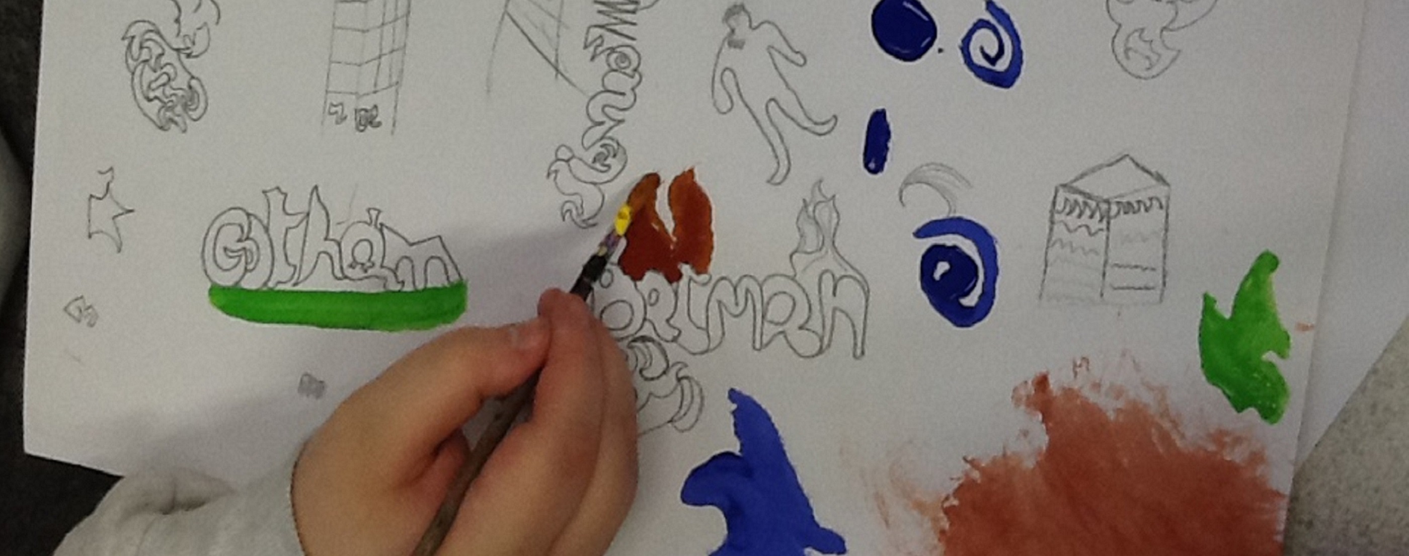 A hand holding a paintbrush painting brown onto a piece of paper to look like flames coming off of the outlined word 'Batman'. Also on the page are other words such as 'Gotham', line drawings of people and building, blue swirls and splashes of green and brown.