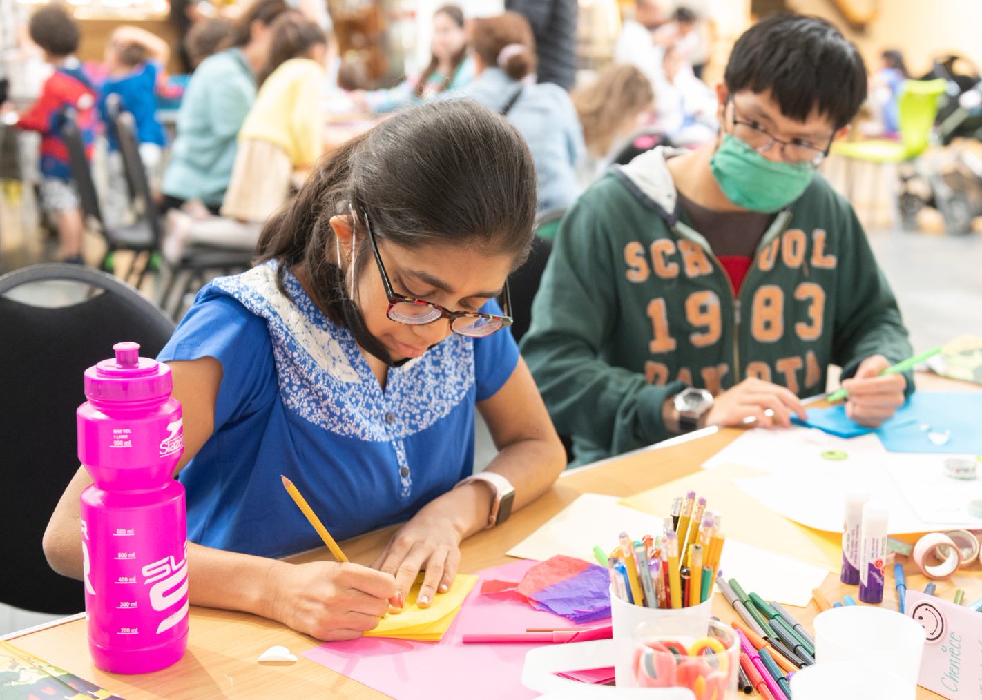 Two young people, Davina and Gar Wa, sat at a table covered in paper and pencils. They are making a zine as part of a community festival at artsdepot, which they helped out with as part of their SEN work experience.  